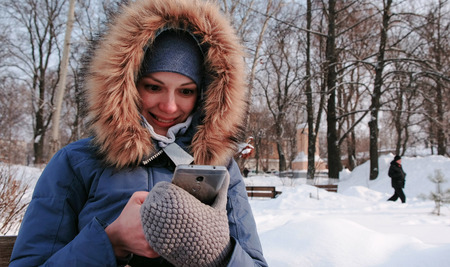 Smiling woman is sitting on bench and browsing mobile phone in winter park in the city in sunny day.の写真素材