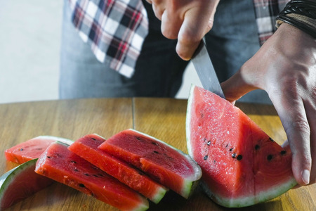 Man cuts watermelon into small slices on the kitchen table. Hands close up.の写真素材