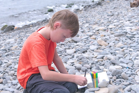 Boy is painting with markers while sitting on a stone beach on the sea shore