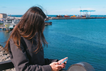 Brunette woman types a message on a mobile phone standing on the waterfront by the sea on a Sunny autumn day.の写真素材