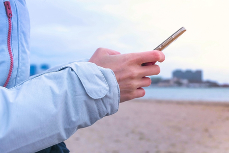 Close-up womans hands typing a messaje on the phone on sea background. Sea winter, woman in white jacket.の写真素材