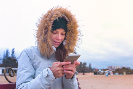 Woman in a white down jacket sitting on a bench on the beach and typing a message on mobile phone.の写真素材