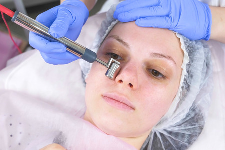 Cosmetologist makes a micro-current procedure for the face of a young woman. Face close-up, front...