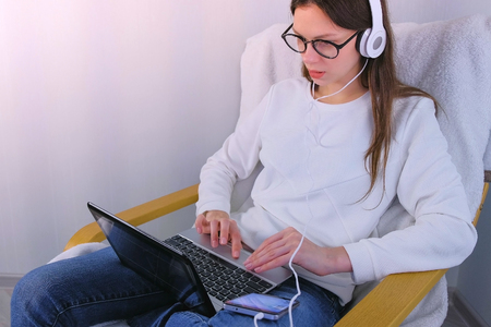 Woman in glasses and headphones is working on laptop sitting in armchair.の写真素材