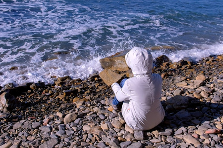 Woman in a white down jacket and hood sits on the shore and looks at the stormy sea waves beating against the stone shore. Back view.の写真素材