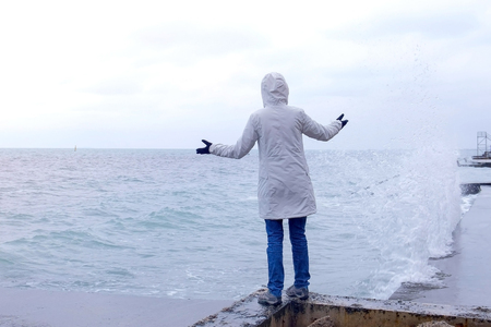 Woman in a white jacket stands on the waterfront on the beach looks at storm waves. Back view.の写真素材