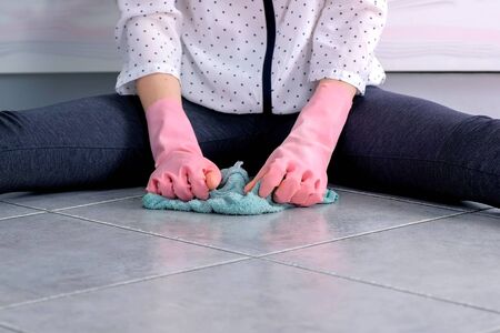Woman in pink rubber gloves washes and rubs hard the stain on kitchen floor with a cloth. Gray tiles on the floor. Hands close-upの写真素材