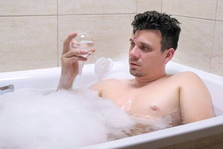 Drunk adult man is falling asleep with wine glass in foam bath at home and drops the glass in the water.の写真素材