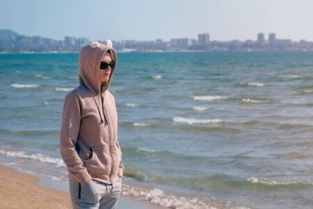 Woman in sweatshirt and sunglasses stands on sand beach on bright Sunny day. Concept of relaxation...