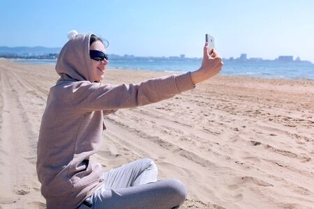 Young 30s woman in sunglasses and hoodie makes selfie on mobile phone, sitting on the beach by sea...