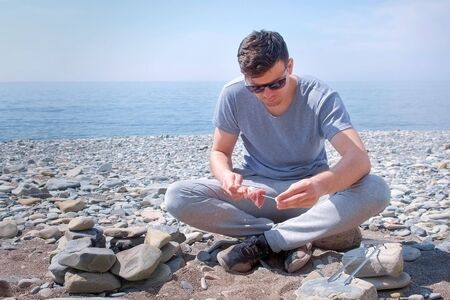 Young man in glasses is preparing shashlik putting the slices of meat on the skewer sitting on sea...