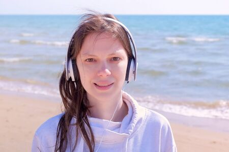 Portrait of young brunette tired woman in headphones on sea background. She is listening music and looking at camera after jogging.の写真素材