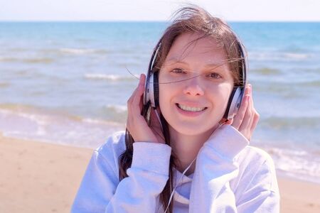 Portrait of young brunette woman in headphones is enjoying music with close eyes. She is walking at sea sand beach.の写真素材
