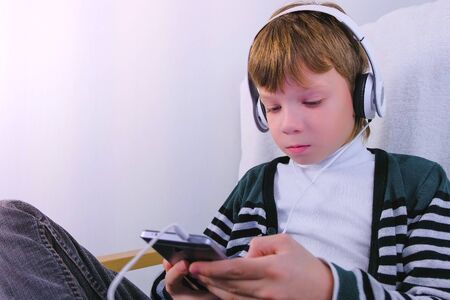 Boy is watching a video in headphones in mobile phone sitting in armchairの写真素材