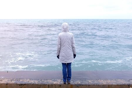 Woman in a white jacket stands on the waterfront on the beach looks at storm waves. Back viewの写真素材