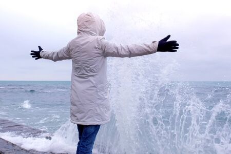 Woman in a white jacket stands on the waterfront on the beach looks at storm waves. Side viewの写真素材
