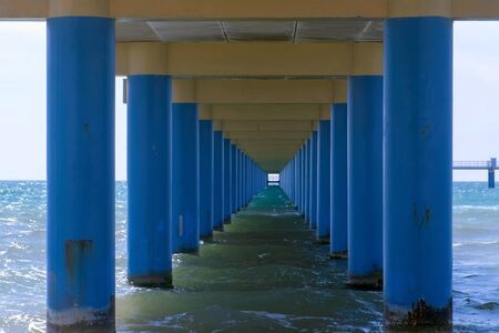 Sea under pier among parallel blue columns making narrow corridor in water. Seascape through pier with sea waves and blue sky at sunny day.の写真素材