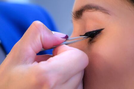 Makeup artist glues artificial eyelashes to girl model in salon, face and eyes closeup, side view. Professional visagiste making extension lashes. Glamour fashion industry. Beauty business concept.の写真素材