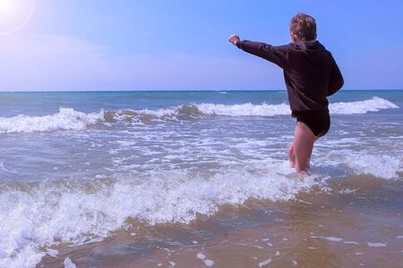 Boy on happy vacations at seashore. Child boy plays and jumps through waves at sea sandy beach in windy spring weather. He wears black sweatshirt and walks at sea water. Tourist travel seaside.の写真素材