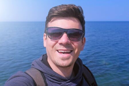 Selfie video of young man tourist blogger talks with subscribers on sea background. He waves hand, greets and smiles. Portrait of guy in sunglasses. Traveller travel journey sea vacation.の写真素材