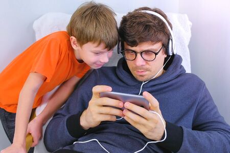 Man in glasses and headphones playing a game in smartphone with his son. Gamer relaxの写真素材