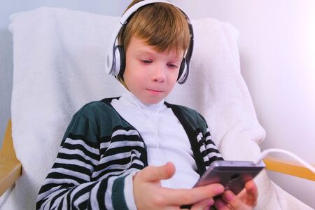 Boy is watching a video in headphones in mobile phone sitting in armchair