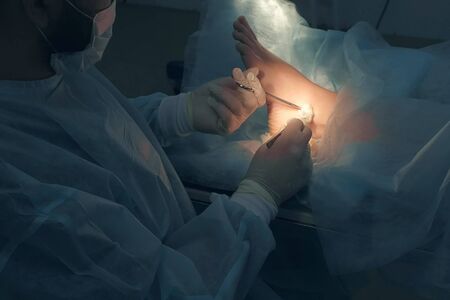 Surgeon man making surgery of removal ankle hygroma in hospital in operating room. Doctor presses cotton swab to patient ankle with clamp. Operation surgical treatment of hygroma on leg.の写真素材