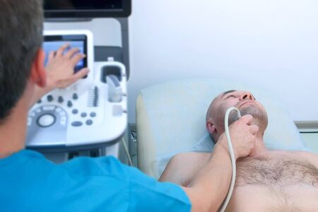 Checkup and scanning of thyroid gland at ultrasound device scanner machine in hospital for man. Doctor runs ultrasound sensor over patients neck and looking at screen. Examination for diagnostic.の写真素材