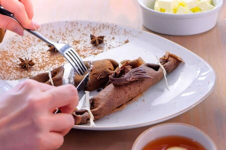 Woman, is eating chocolate russian pancakes, blini with curd filling on the plate. Hands close-up. Served with tea. Close-up hands, side viewの写真素材