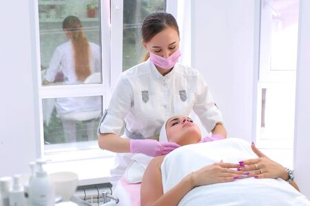 Cosmetologist in gloves washes cleans clients woman face wiping with napkins. Beautician making massaging beauty facial skincare procedure to patient in clinic. Beauty industry concept.の写真素材