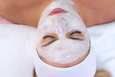 Portrait of woman laying in spa salon with white mask on face, top view. Beauty facial skincare procedure to patient in cosmetology clinic, face closeup. Beauty industry concept.の写真素材