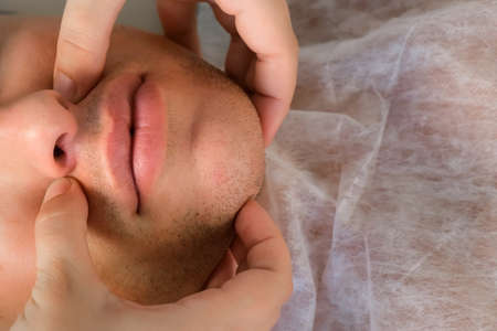 Manual therapist doctor is making facial massage for young man, closeup face top view. Treatment of headaches with physiotherapist. She presses her fingers on certain points on the patients neck.の写真素材