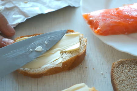 Man spreads butter on a piece of wheat bread using knife doing sandwich with red fish for breakfast, hands closeup. Preparing food at home concept. Fast food nutrition.の写真素材