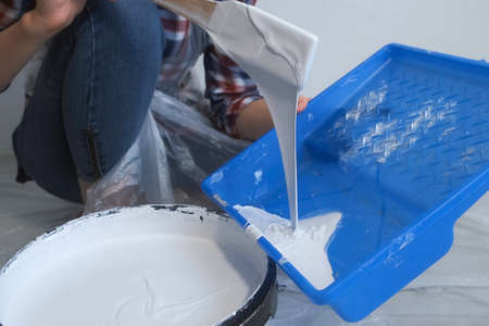 Woman painter is pouring paint in a tray for painting walls using wooden spatula, closeup view. DIY...