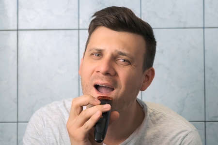 Handsome young man is shaving beard and moustache using electric razor, front view. He is looking at...