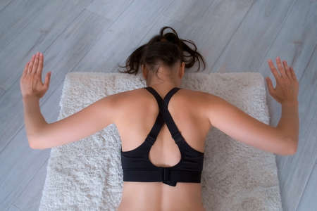 Fit slim young woman is training on fitness workout at home lying on carpet, back view. She is doing physical exercises for shoulders lifting arms from floor. Sport, wellness concept, losing weight.の写真素材