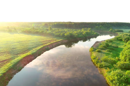 Flying over river and green forest at summer sunny day, sky reflecting in water. Sunrise on nature in beautiful place with natural landscape. Aerial view of picturesque woodland and river.の写真素材