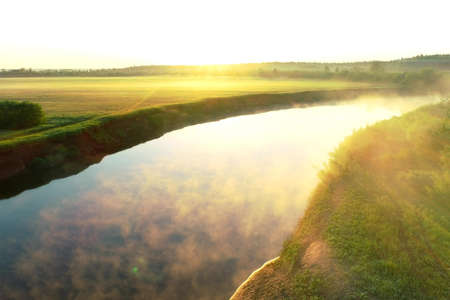 Flying over river with white mist and green forest at summer sunny day, sky reflecting in water. Sunrise on nature in beautiful place with natural landscape. Aerial view of picturesque woodland.の写真素材
