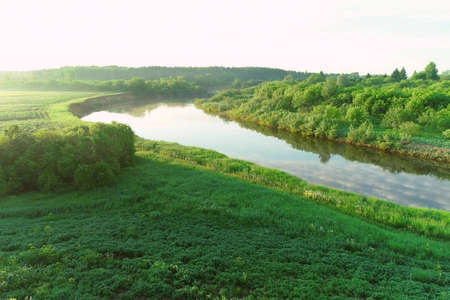 Flying over river and green forest at summer sunny day, sky reflecting in water. Sunrise on nature in beautiful place with natural landscape. Aerial view of picturesque woodland and river.の写真素材