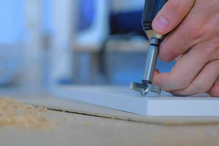 Man drills a round hole in the chipboard making furniture wardrobe door, closeup side view. Guy is drilling a hole for the door hinges, hands closeup. Doing renovation at home by himself.の写真素材