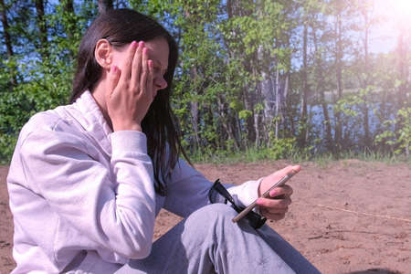 Woman brushes off midges sitting in forest by lake reading smartphone, side view. Brunette young...