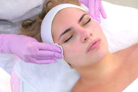 Cosmetologist in gloves cleans clients woman face, neck and chest wiping with cotton pads....