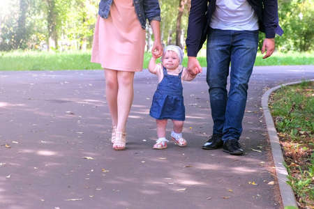 Mother and Father learning their daugter to walk outdoors. Baby girl is trying to go making first steps holding mom and dads hands in city park. Happy girl of one year. Parenthood babycare concept.の写真素材
