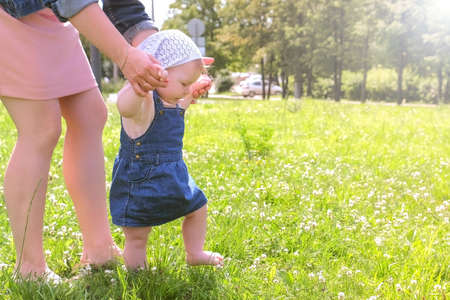 Mom learning baby daughter to walk, first steps in park at summer warm day with barefooted legs in...