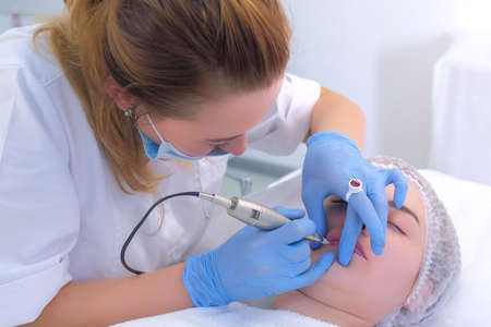 Beautician applying permanent makeup on woman lips drawing using tattoo machine and pink pigment....