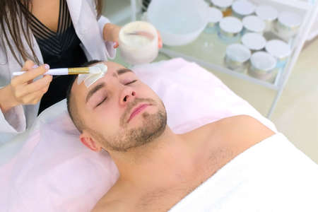 Cosmetologist is applying white moisturizing mask on mans face using brush in beauty clinic, face closeup. Beauty industry concept. Guy is lying on couch. Beautician woman making spa procedure.の写真素材