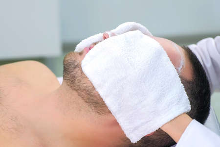 Cosmetologist wipes white moisturizing mask from mans face using terry mittens in beauty clinic, side view, face closeup. Beauty industry concept. Guy is lying on couch. Beautician making procedure.の写真素材