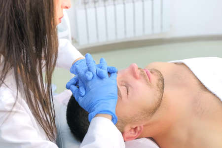 Cosmetologist in gloves is applying cream on clients man face massaging skin, side view. Beautician...
