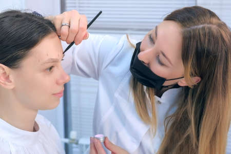Cosmetologist painting brush shape of eyebrows woman before tint procedure in beuty clinic with white paint. Professional beautician at work shaping eyebrows to young girl. Portrait of woman.の写真素材
