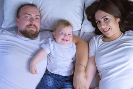 Family portrait of mom, dad and baby lying in bed at home looking at camera and then kissing baby together, top view. Family look style, lovely moments. Happy relationships of young couple.の写真素材